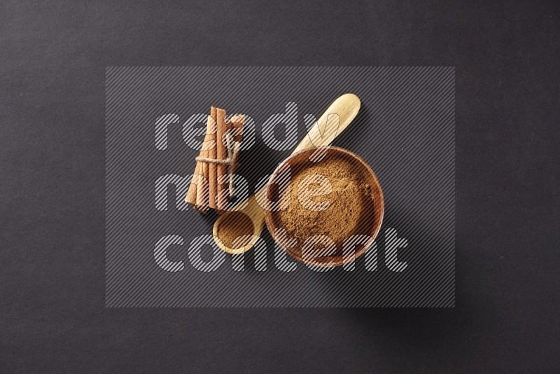 أعواد القرفة مربوطة بجانب وعاء خشبي ممتلئ ببودرة القرفة وملعقة خشبية ممتلئة بالبودرة على خلفية سوداء