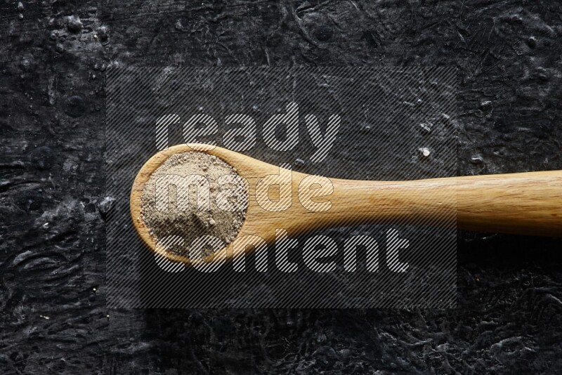 A wooden spoon full of white pepper powder on textured black flooring
