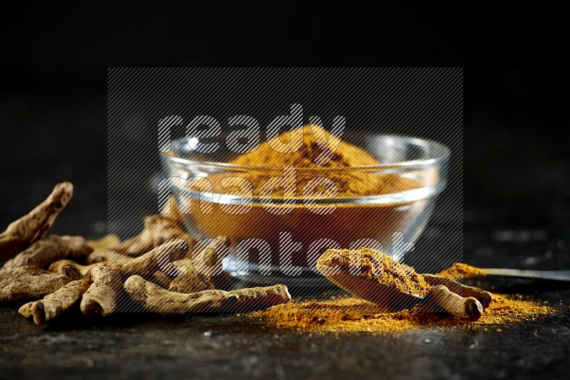 A glass bowl and a metal spoon full of turmeric powder and dried whole fingers next of them on textured black flooring