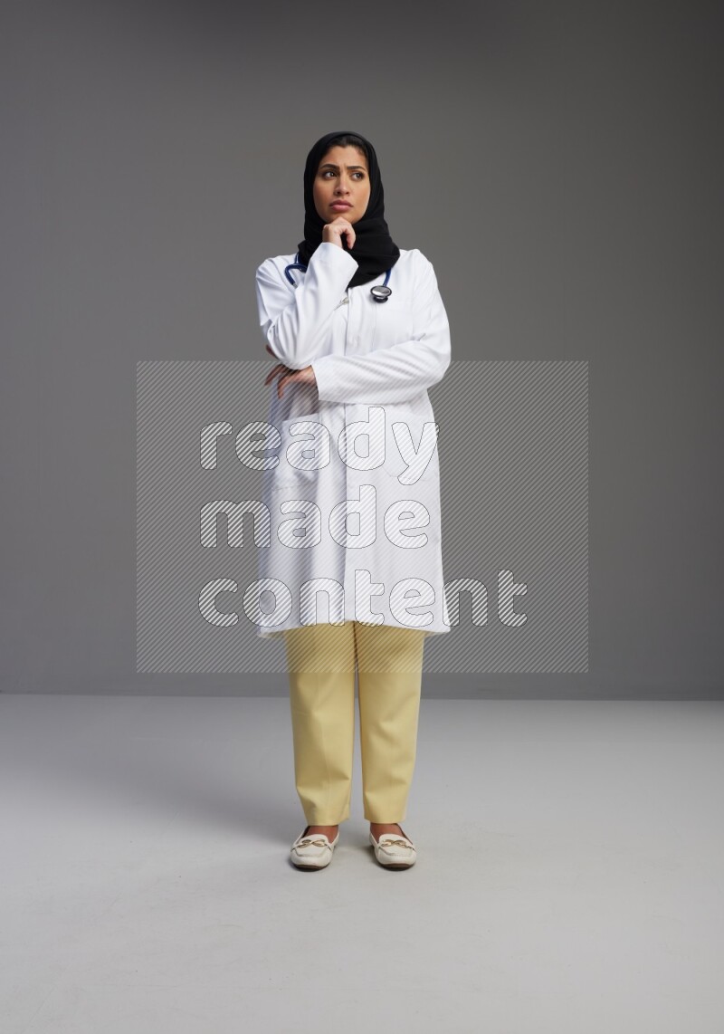 Saudi woman wearing lab coat with stethoscope standing interacting with the camera on Gray background