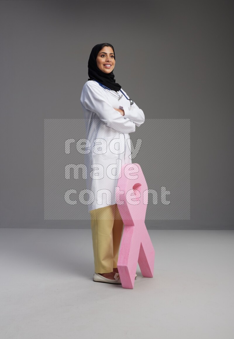 Saudi woman wearing lab coat with stethoscope standing holding awareness ribbon symbol standing on Gray background