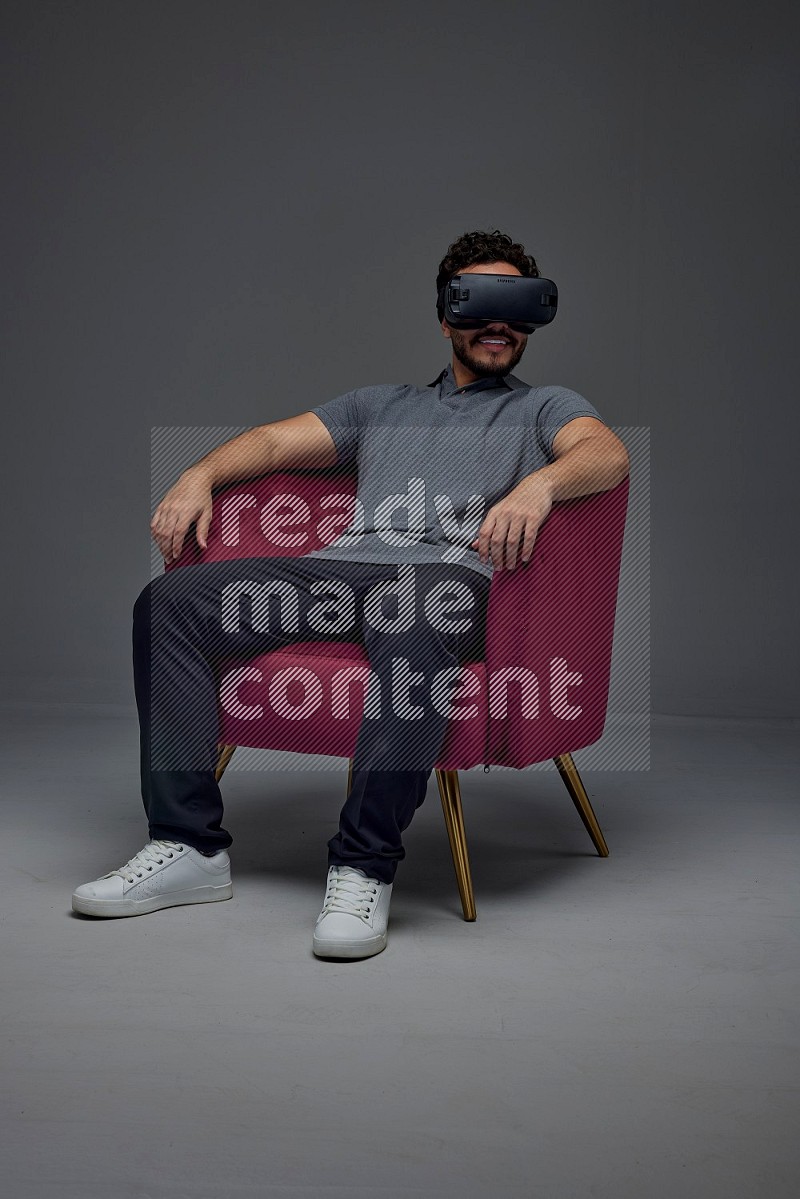 A man wearing casual and wearing VR while sitting on a burgundy chair eye level on a gray background
