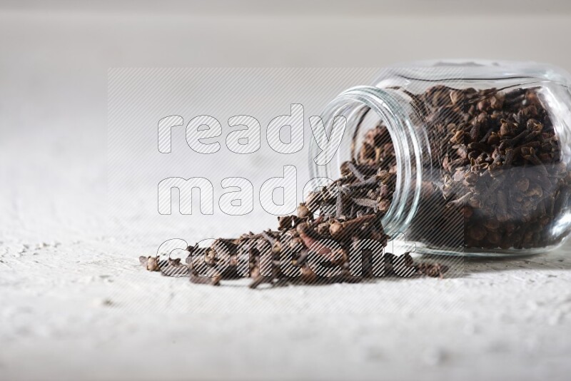A flipped glass spice jar full of cloves and cloves came out of it on textured white flooring