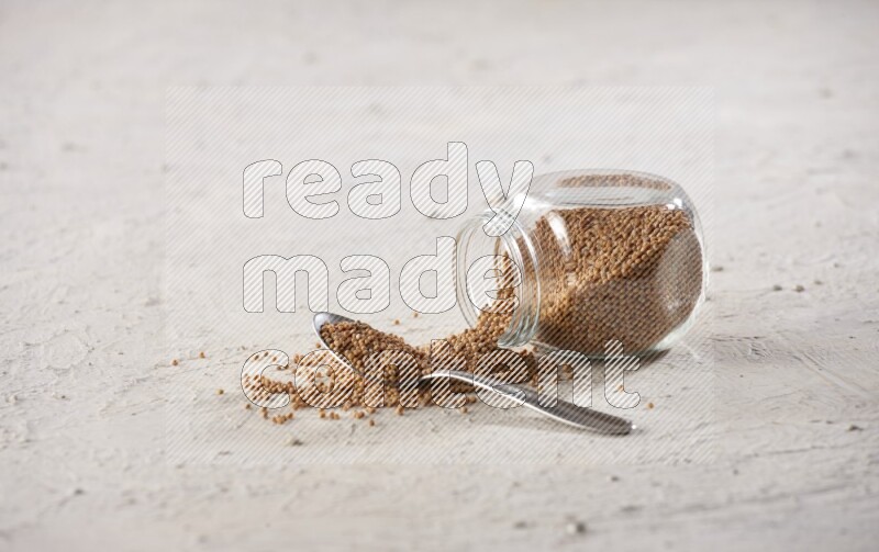A glass spice jar and a metal spoon full of mustard seeds and jar is flipped with fallen seeds on a textured white flooring