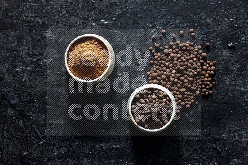 2 beige bowls, one full of allspice powder and the other full of whole balls and both spread on a textured black flooring