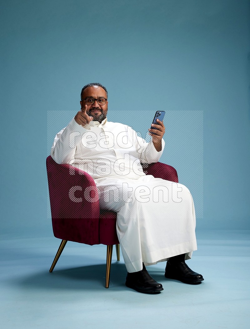 Saudi Man without shimag sitting on chair texting on phone on blue background