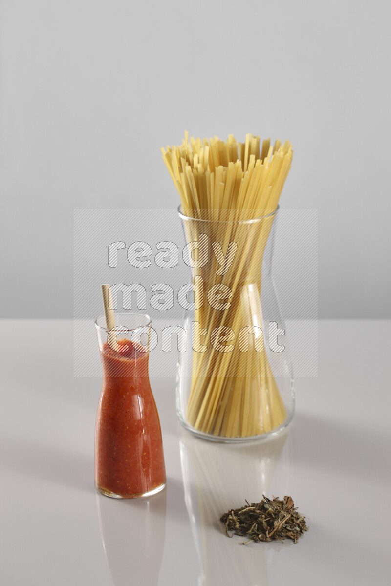 Raw pasta with tomatoe pasta with different ingredients such as cherry tomatoes, basil, garlic, bay laurel, cardamom, white pepper, black pepper, red chilis and wheat stalks on light grey background