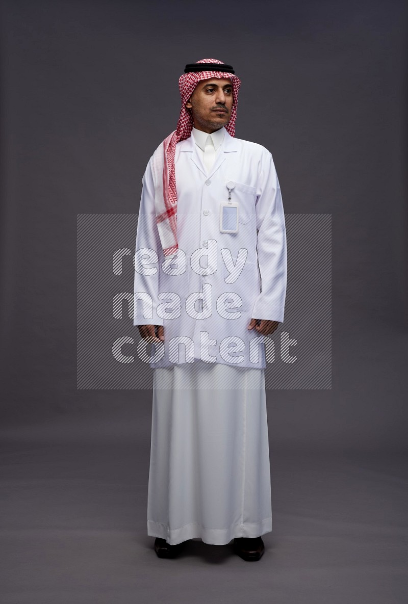 Saudi man wearing thob with lab coat and shomag with pocket employee badge standing interacting with the camera on gray background