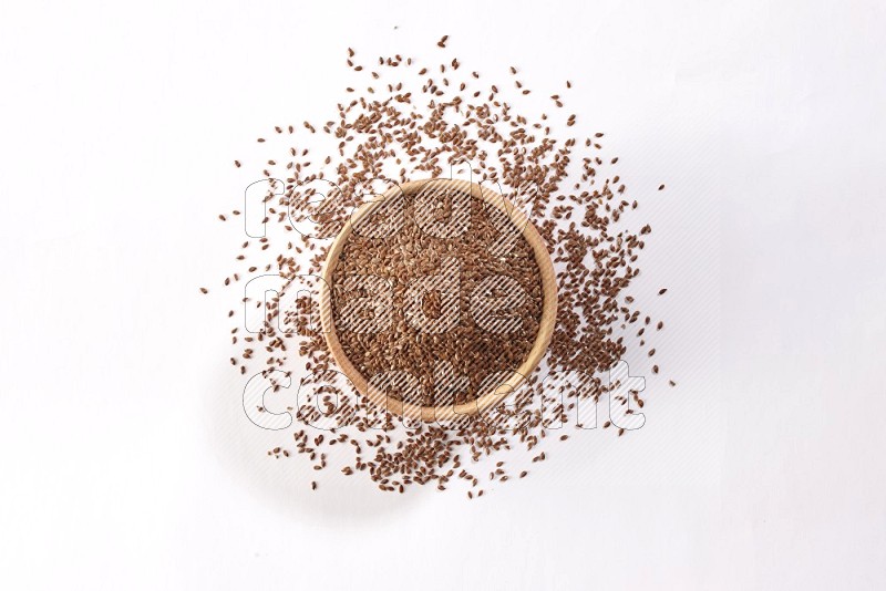 A wooden bowl full of flax seeds surrounded by flax seeds on a white flooring