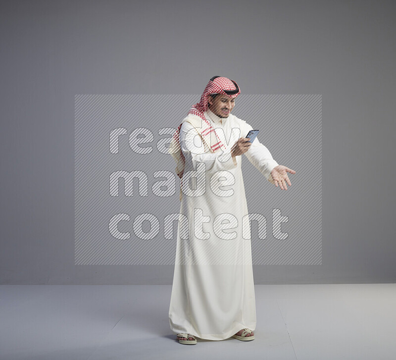 A  Saudi man standing wearing thob and red shomag texting on phone on gray background