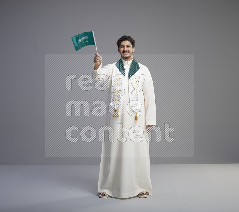 A Saudi man standing wearing thob and saudi flag scarf and holding small saudi flag on gray background