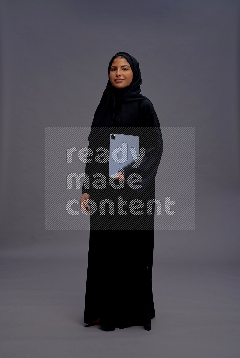 Saudi woman wearing Abaya standing working on tablet on gray background