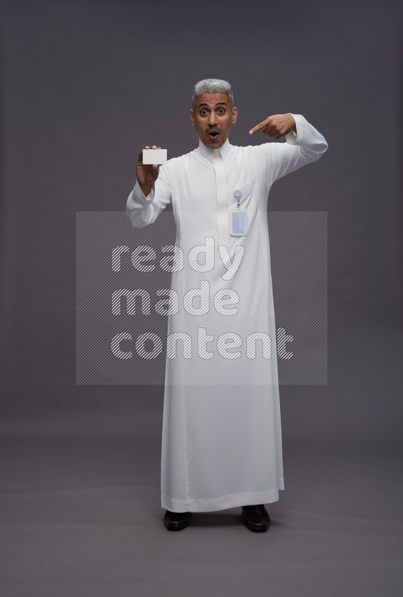 Saudi man wearing thob with pocket employee badge standing holding ATM card on gray background