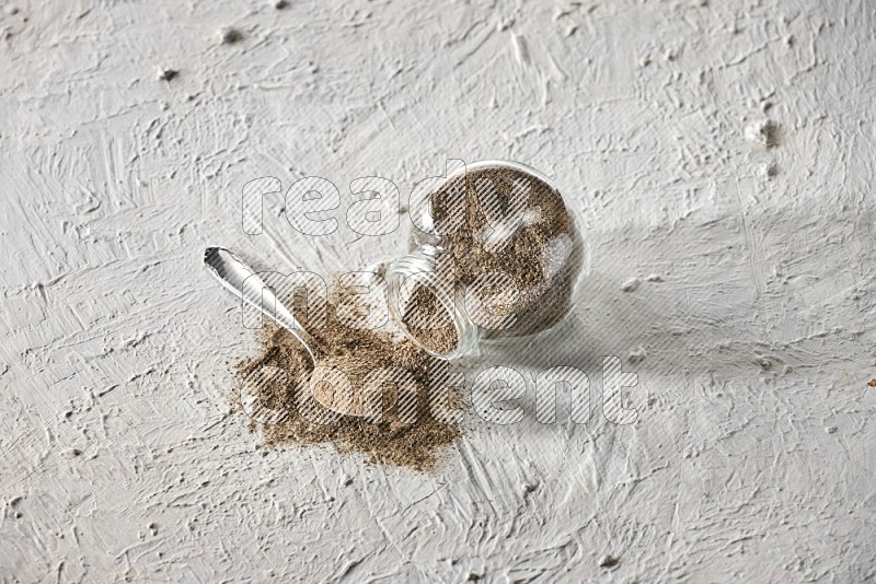 Flipped glass spice jar full of black pepper powder with a metal spoon full of it on textured white flooring