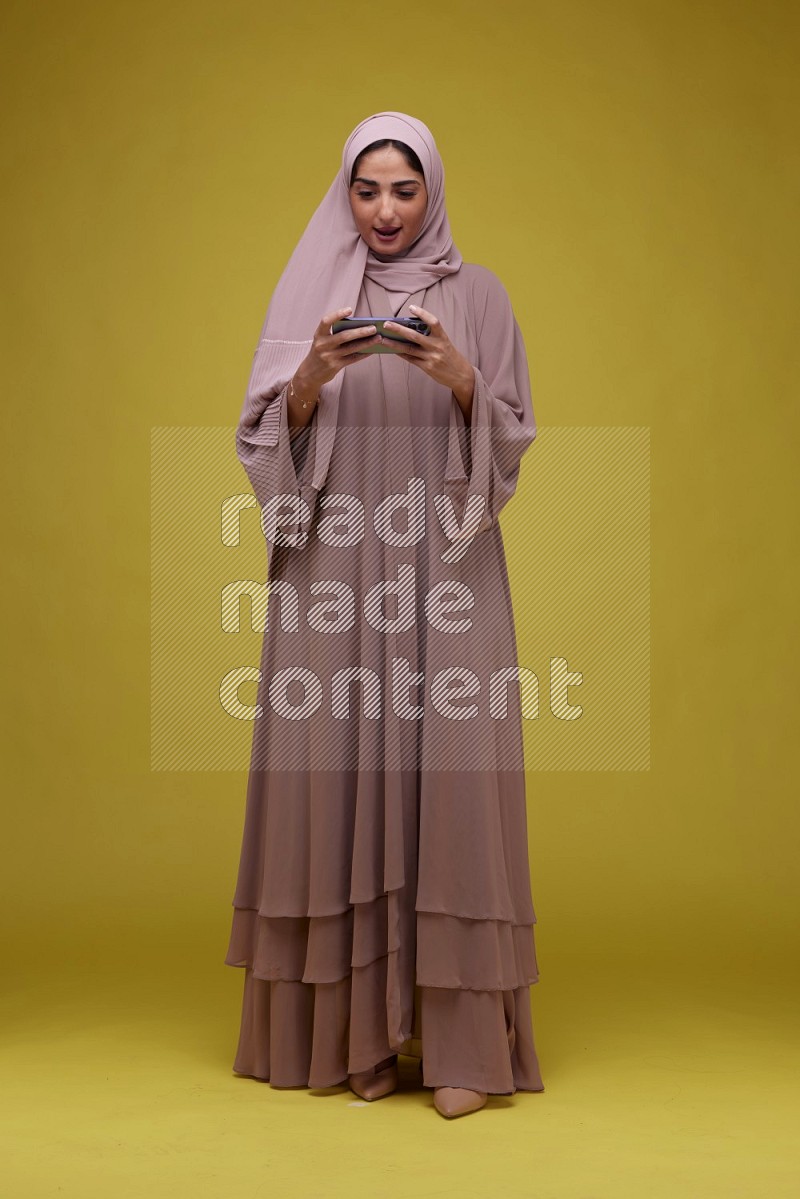 A woman Playing Games a Yellow Background wearing Brown Abaya with Hijab
