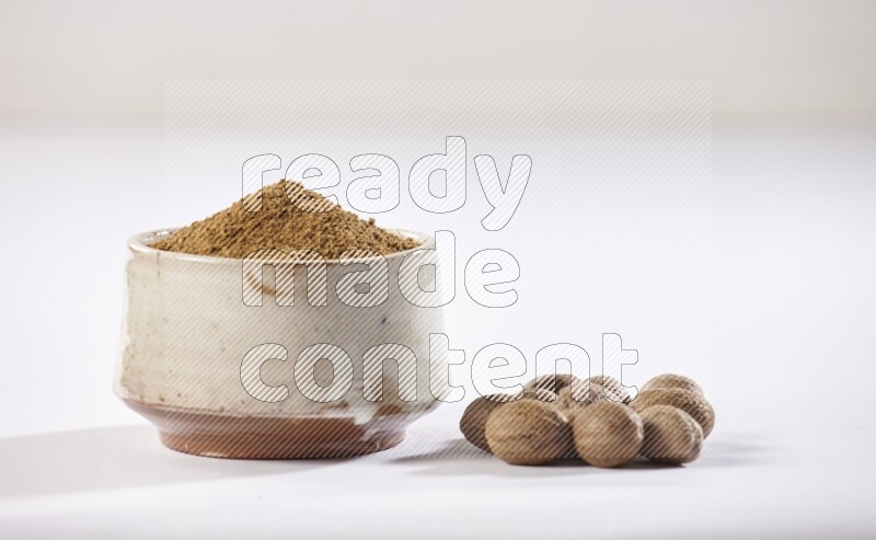 A beige pottery bowl full of nutmeg powder with whole seeds beside it on a white flooring