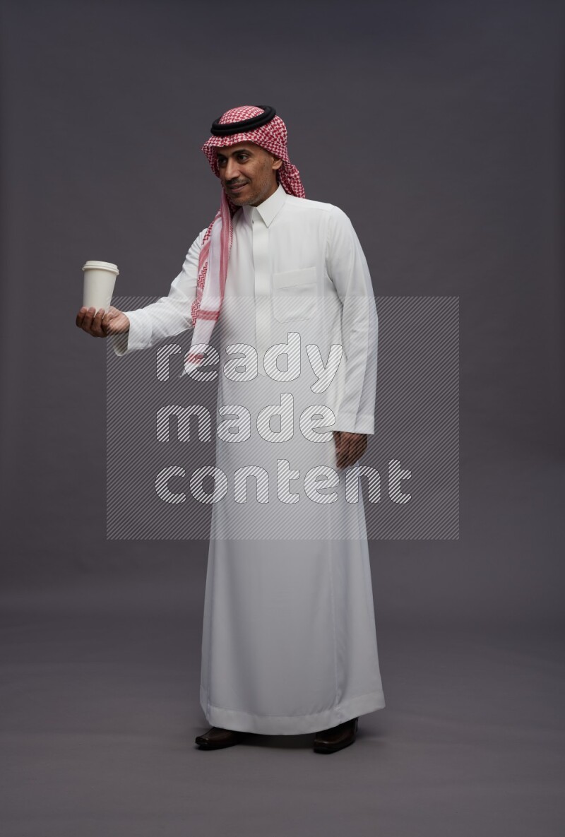Saudi man wearing thob and shomag standing holding paper cup on gray background