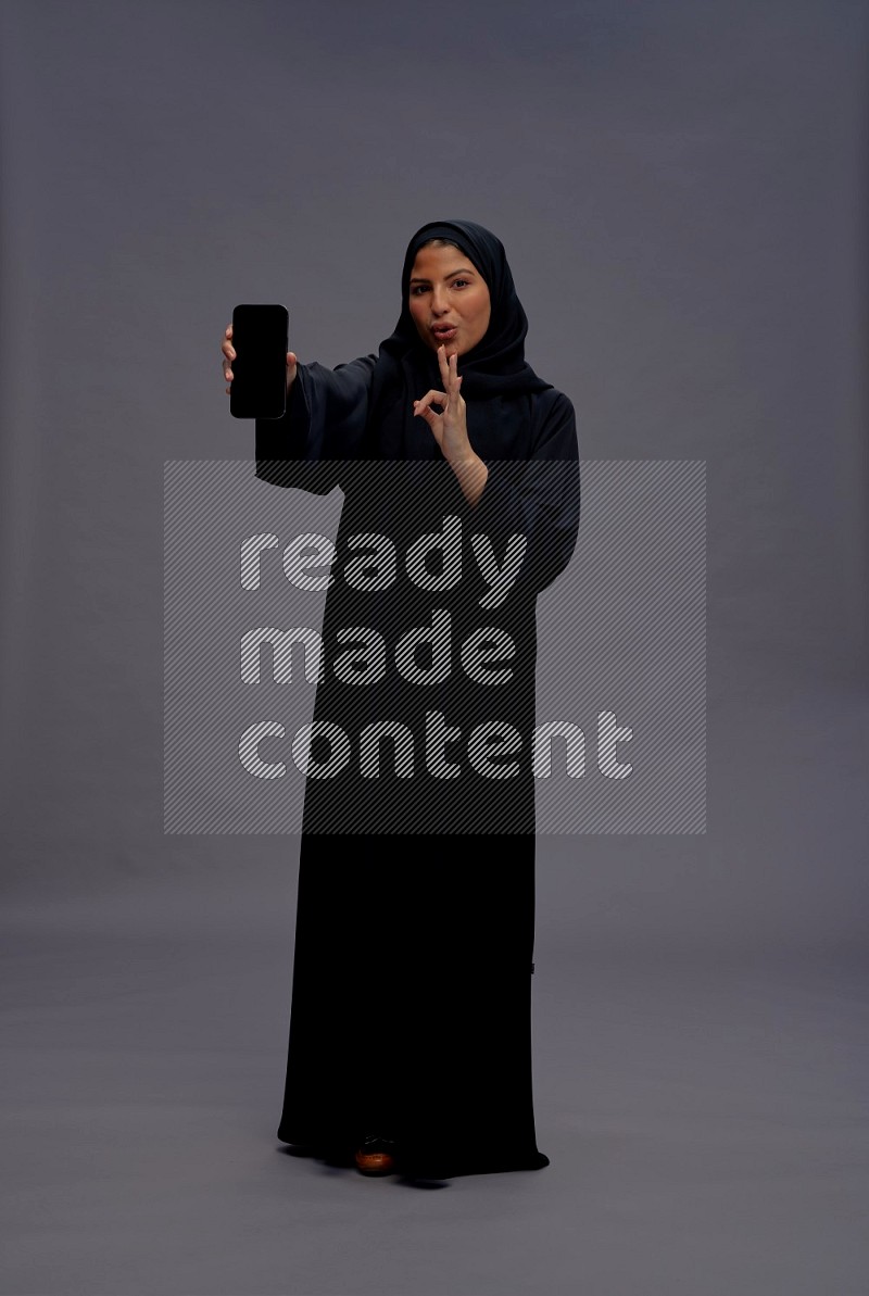 Saudi woman wearing Abaya standing showing phone to camera on gray background