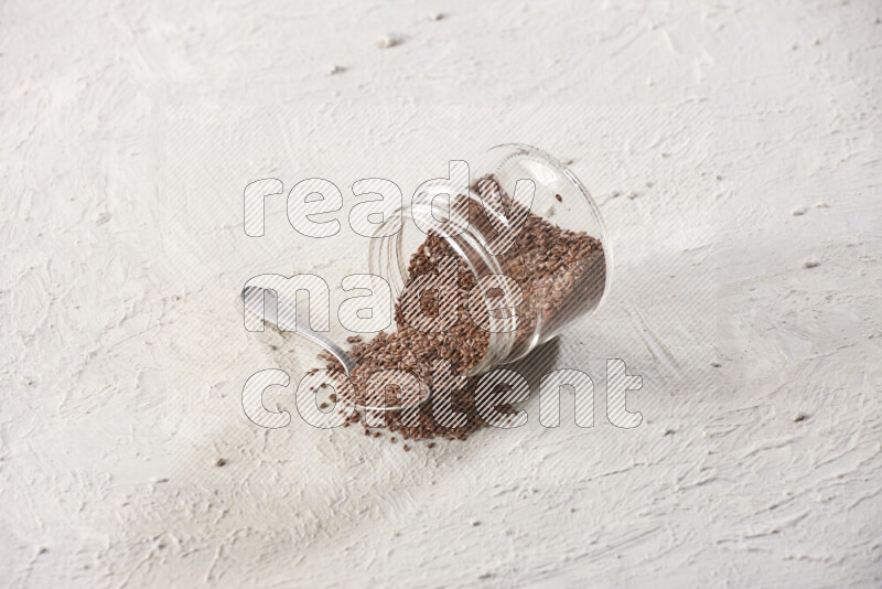 وعاء زجاجي مقلوب ممتلئ بحبوب بذر الكتان مع ملعقة معدنية ممتلئة علي خلفية بيضاء