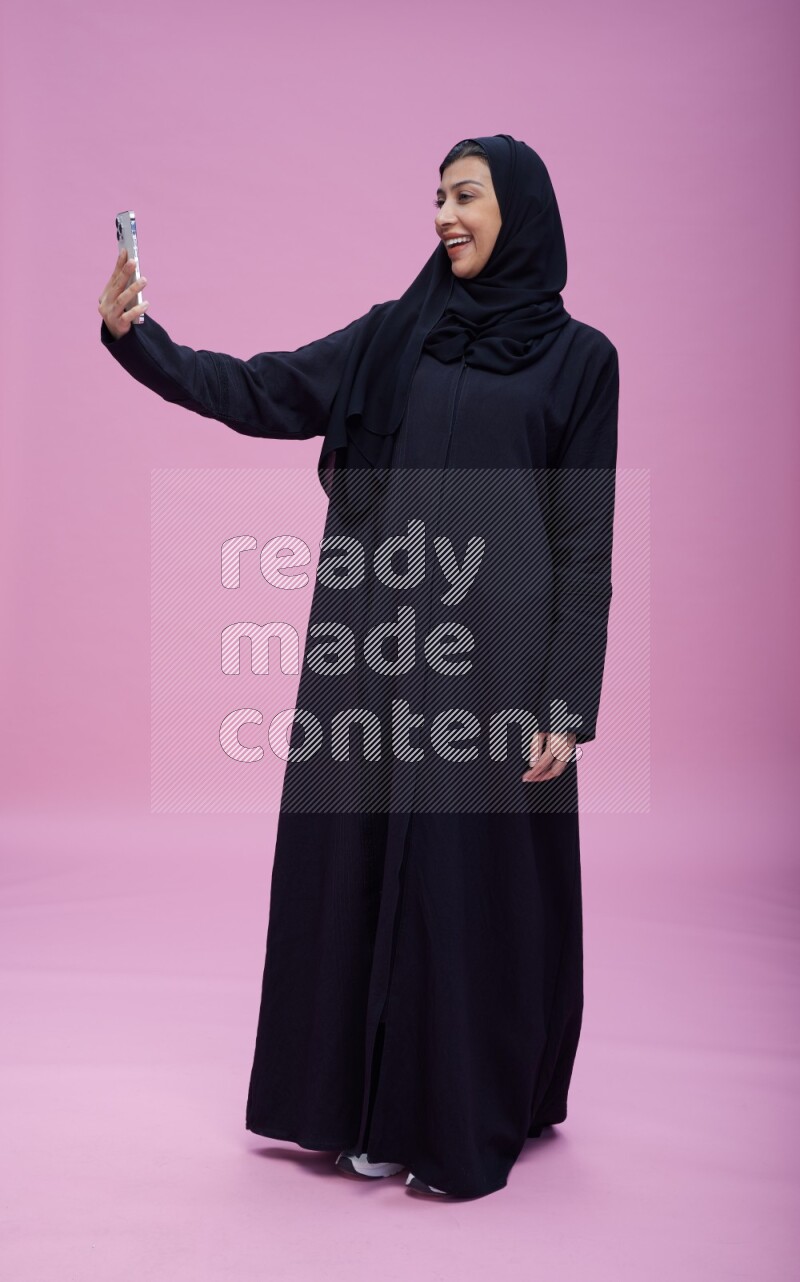Saudi woman wearing Abaya standing taking selfie on pink background
