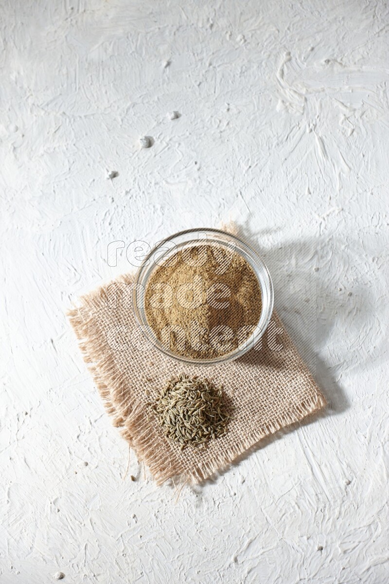 A glass bowl full of cumin powder with some of cumin seeds on burlap piece on a textured white flooring