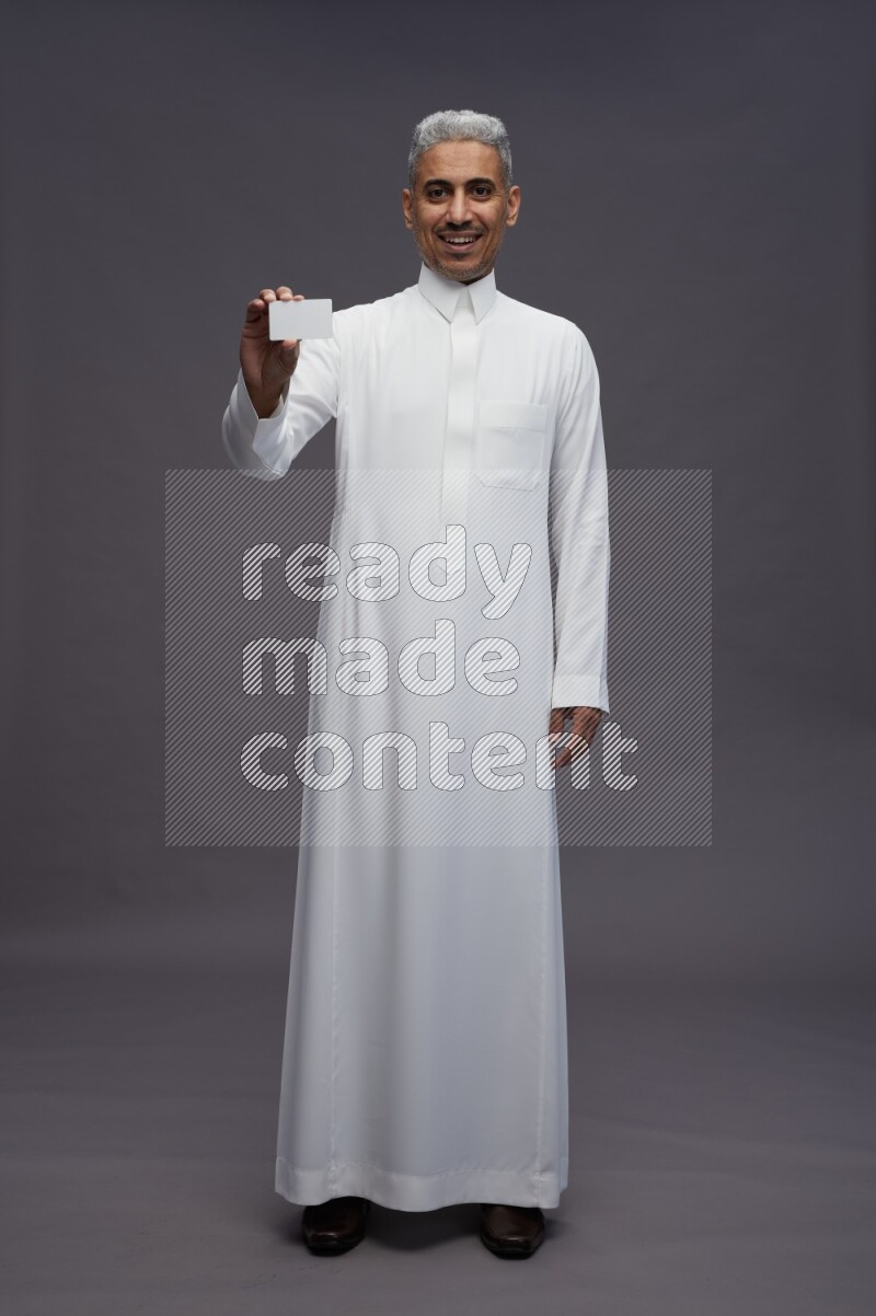 Saudi man wearing thob standing holding ATM card on gray background