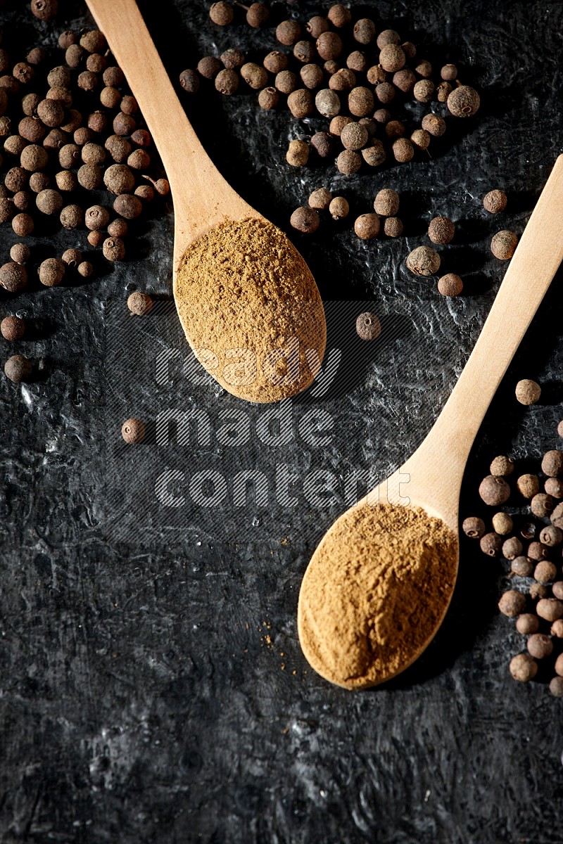 Wooden spoons full of all spice powder and allspice whole balls beside it on a textured black flooring