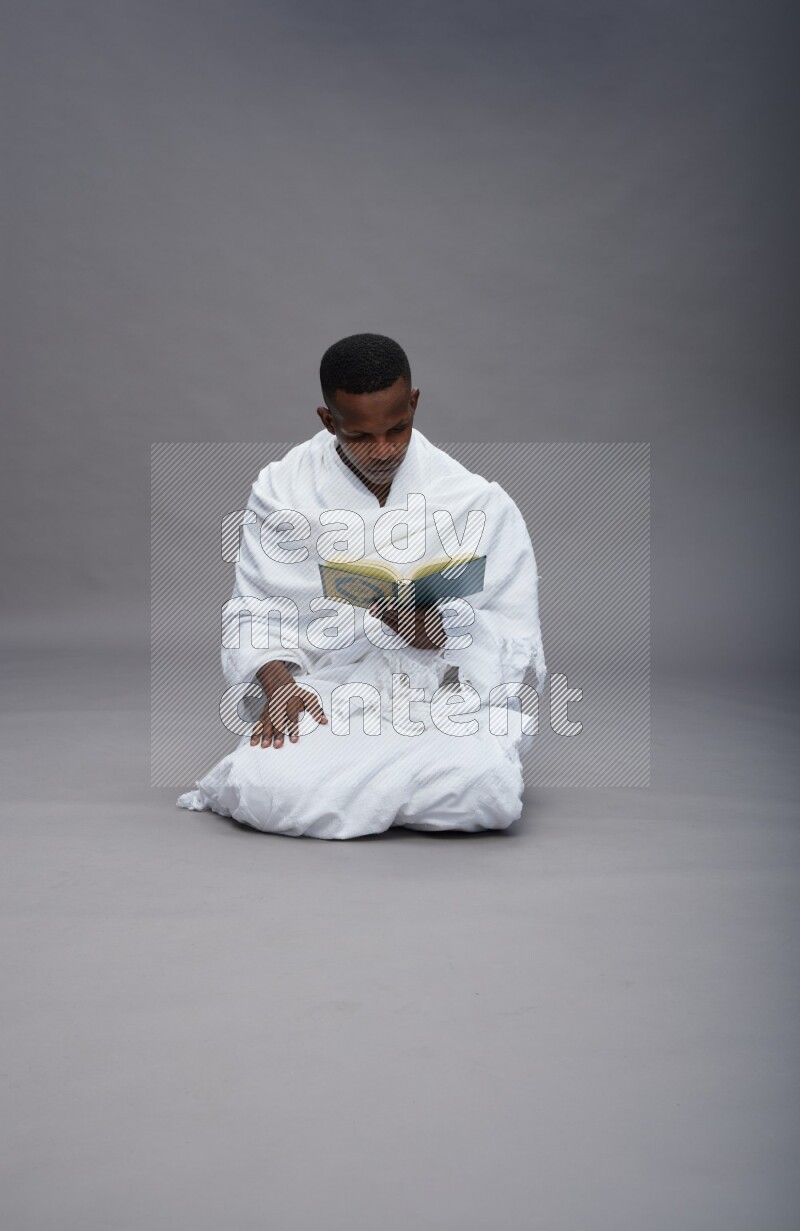 A man wearing Ehram sitting on floor reading quran on gray background