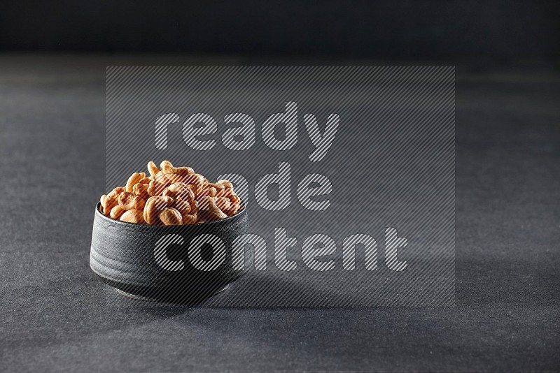 A black pottery bowl full of cashews on a black background in different angles