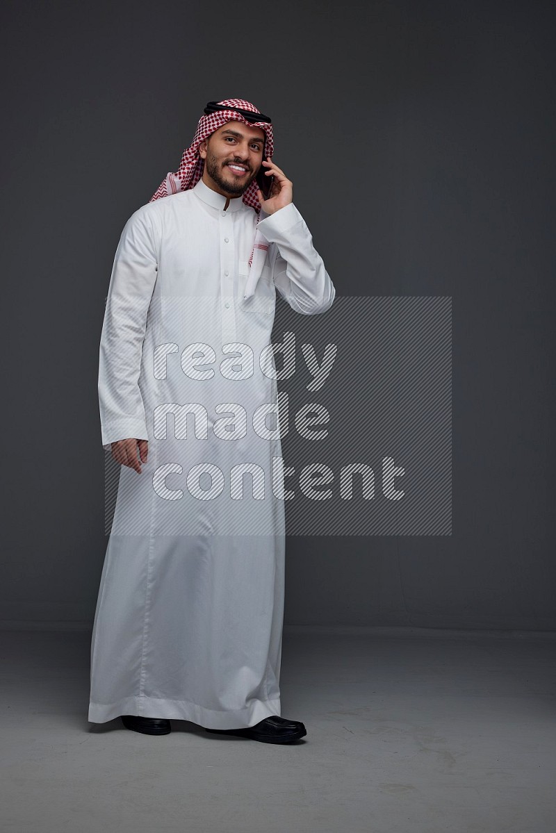 A Saudi man wearing Thobe and Shmagh talking in the phone eye level on a gray background