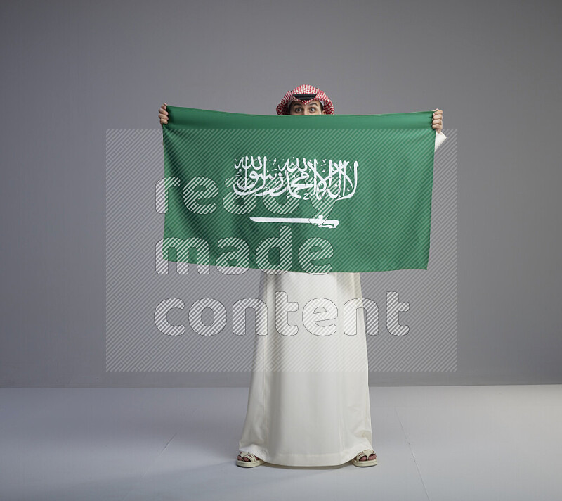 A saudi man standing wearing thob and red shomag holding big saudi flag on gray background