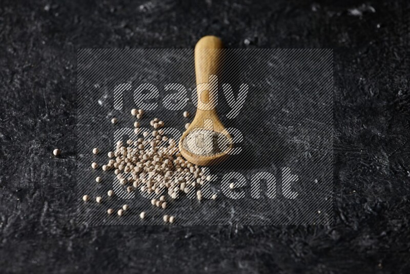 A wooden spoon full of white pepper powder with white pepper beads on textured black flooring