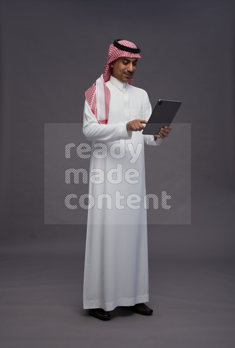 Saudi man wearing thob and shomag standing working on tablet on gray background