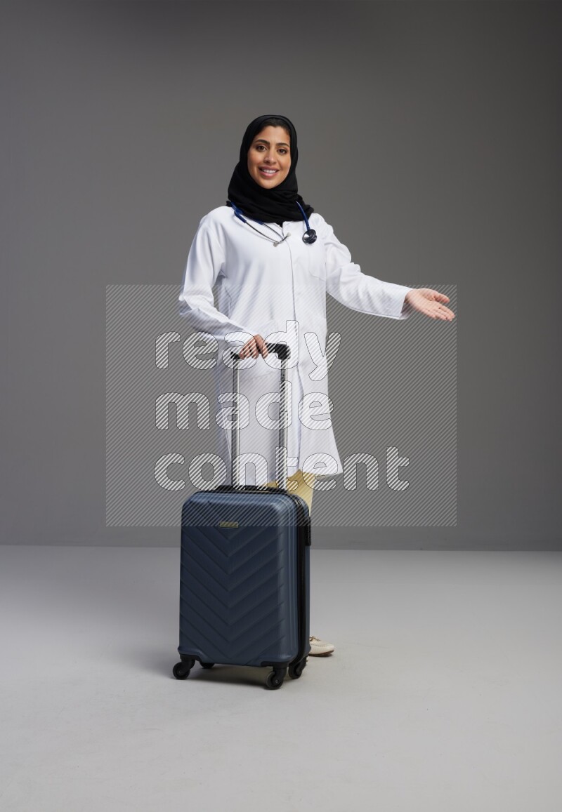Saudi woman wearing lab coat with stethoscope standing holding Travel bag on Gray background