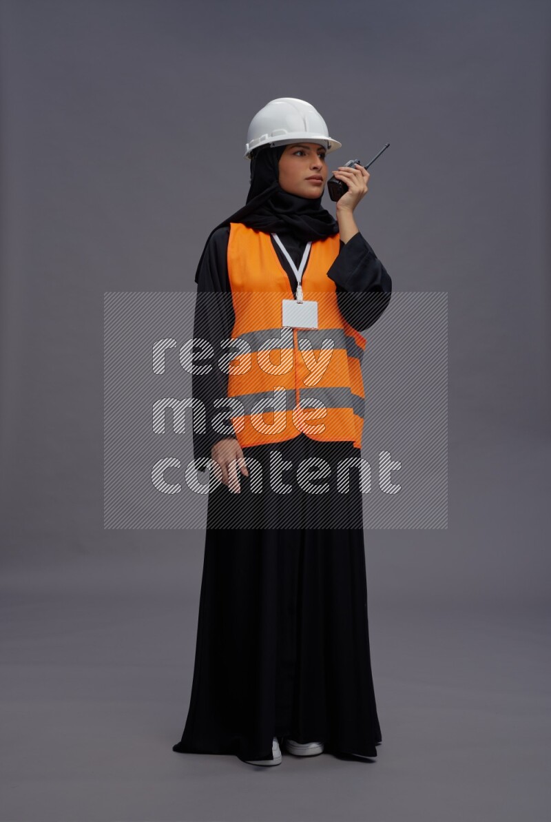 Saudi woman wearing Abaya with engineer vest with neck strap employee badge standing holding walkie-talkie on gray background