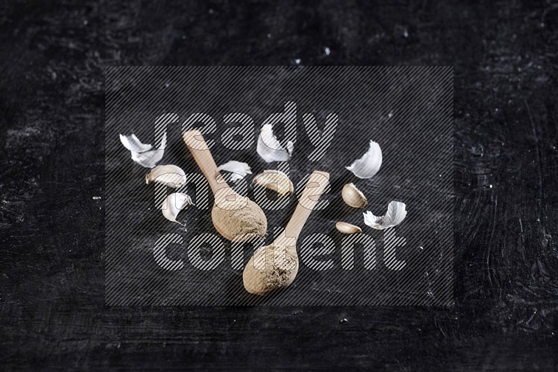 Two wooden spoons filled with garlic powder, surrounded by peeled garlic cloves and their skins on a textured black flooring