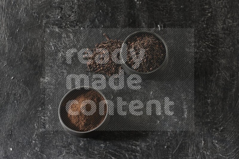 2 Black pottery bowls full of cloves and the other full of cloves powder on textured black flooring