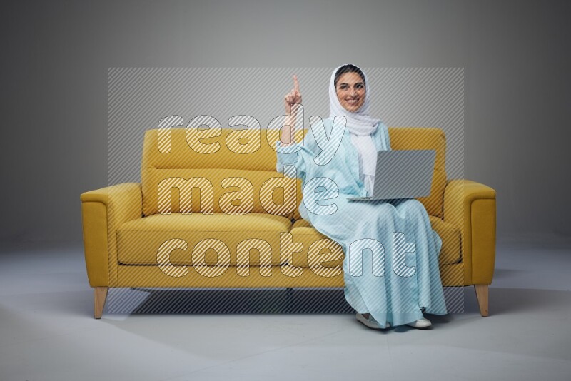 A Saudi woman sitting on a yellow Sofa With her Laptop Pointing wearing Blue Abaya