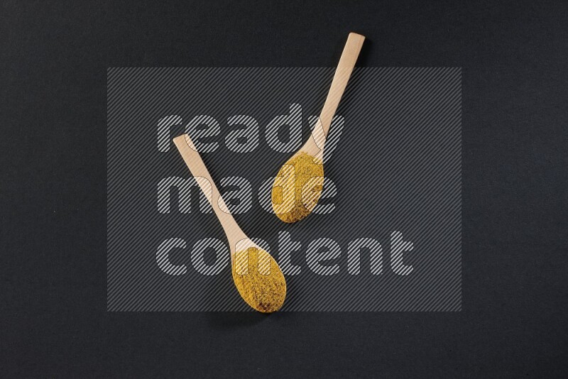 2 wooden spoons full of turmeric powder on black flooring
