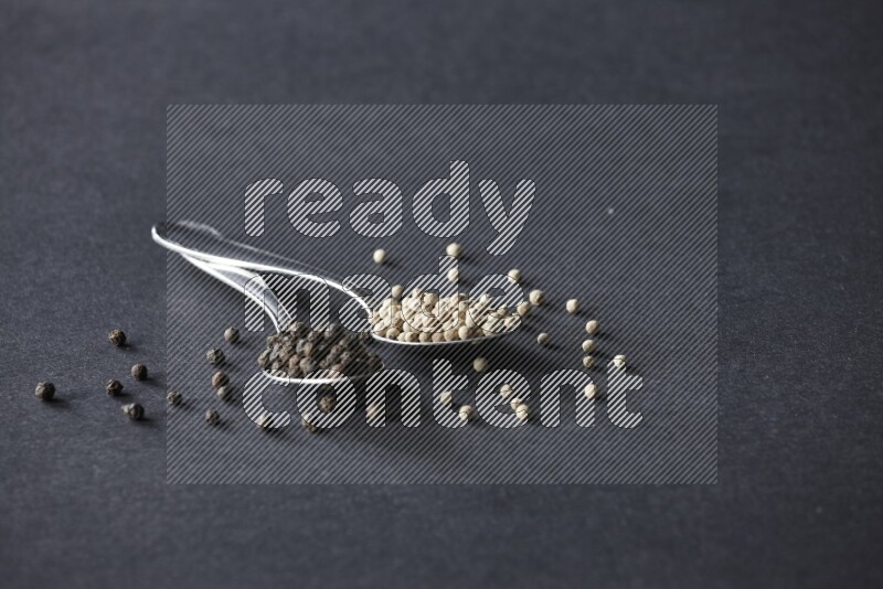 2 metal spoons full of black and white pepper beads with spreaded beads on black flooring