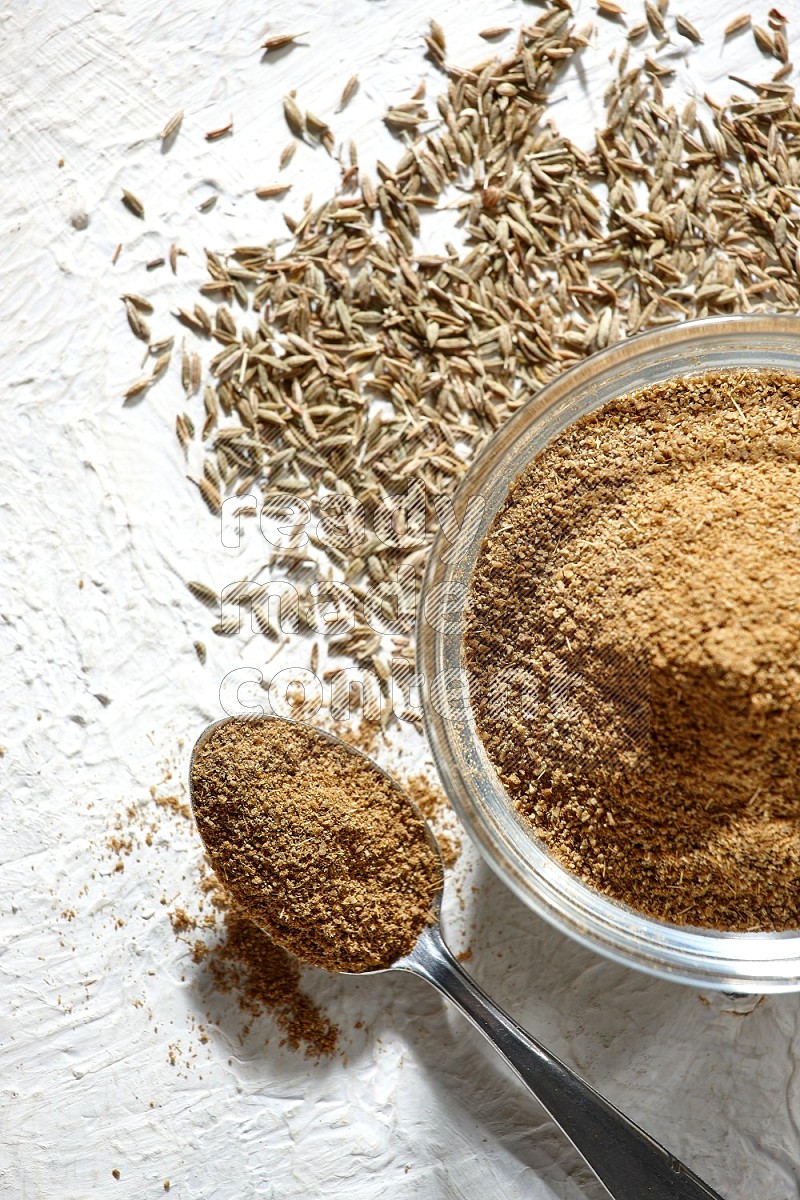 A glass bowl and metal spoon full of cumin powder and cumin seeds underneath it on textured white flooring