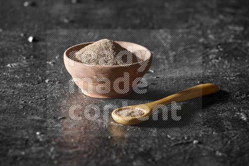 A wooden bowl and wooden spoon full of black pepper powder on a textured black flooring