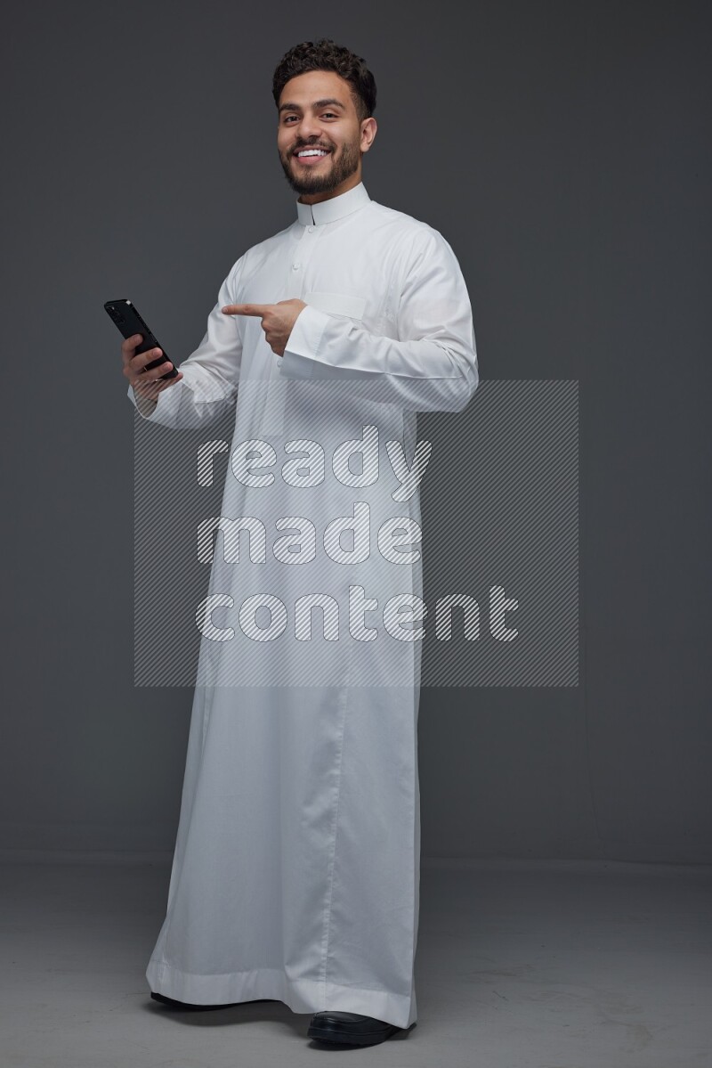 A Saudi man wearing Thobe and holding his phone while pointing to it eye level on a gray background