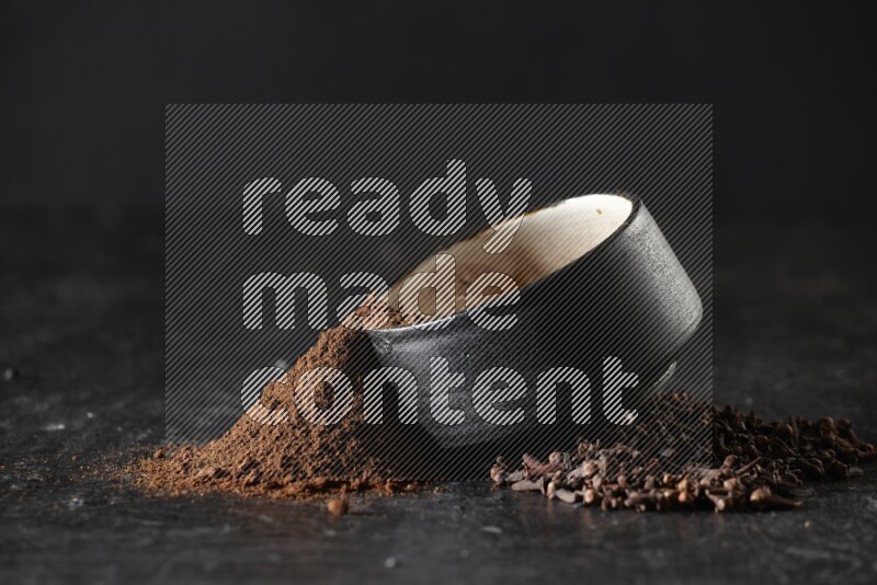 A black pottery bowl full of cloves powder spilled out of it with whole grains on a textured black flooring