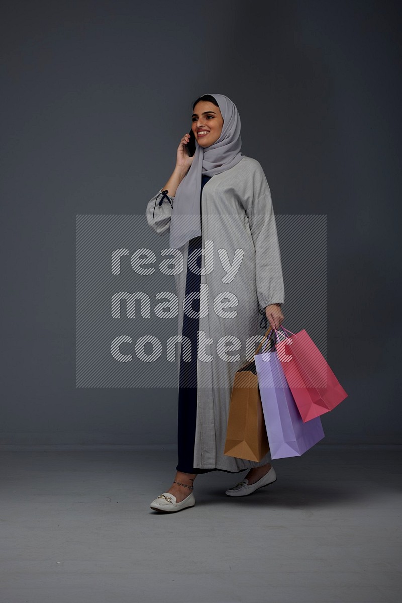 A Saudi woman wearing a light gray Abaya and head scarf standing and holding shopping bags making different poses eye level on a grey background