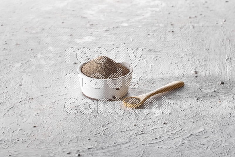 A pottery white bowl full of black pepper powder and wooden spoon full of powder on textured white flooring