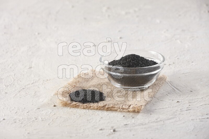 A glass bowl full of black seeds on a burlap piece on textured white flooring