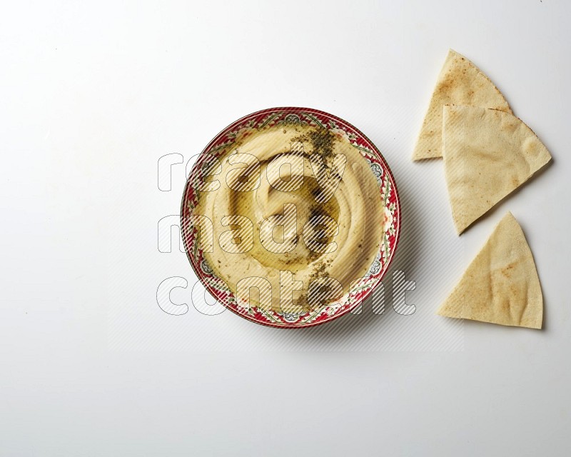 Hummus in a red plate with patterns garnished with zaatar & sumak on a white background