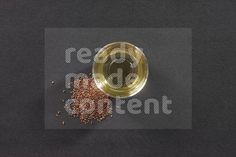 A glass bowl full of flaxseeds oil with some of flaxseeds beside it on a black flooring