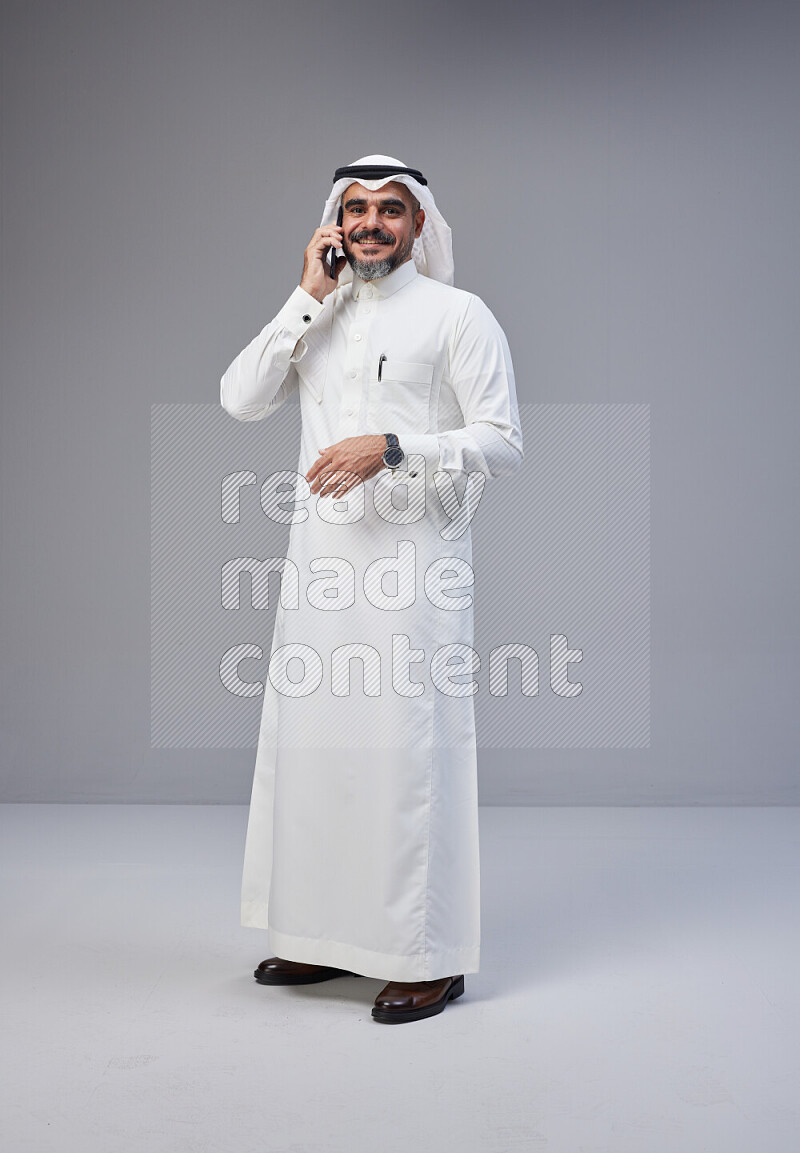 Saudi man Wearing Thob and white Shomag standing talking on phone on Gray background